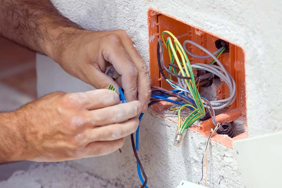 &Eacute;lectricien travaillant sur installation &eacute;lectrique murale, manipulant des c&acirc;bles multicolores sortant d'un bo&icirc;tier orange encastr&eacute; dans mur blanc