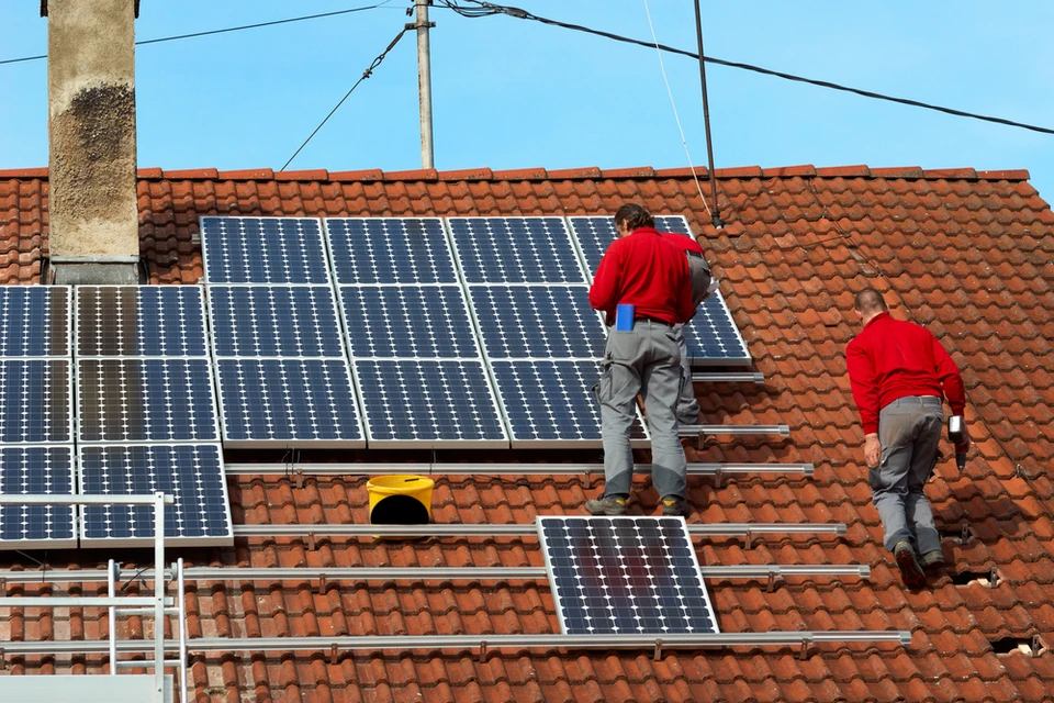 Deux techniciens en uniformes rouges installent des panneaux solaires photovolta&iuml;ques sur une toiture en tuiles rouges d'une maison r&eacute;sidentielle