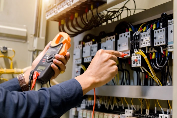 Électricien en uniforme bleu travaillant dans un tableau électrique industriel, utilisant une pince ampèremétrique orange pour mesurer l'intensité électrique