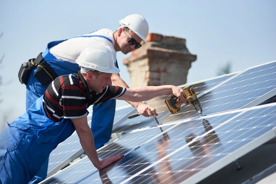Deux techniciens en combinaisons bleues et casques blancs installent des panneaux photovoltaïques sur un toit résidentiel