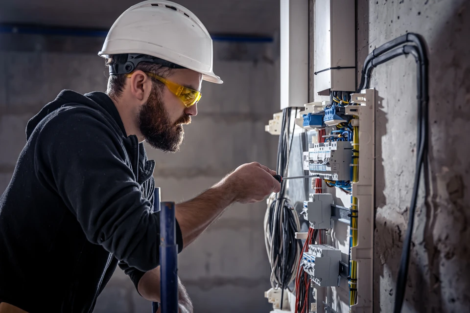 Électricien barbu portant casque blanc et lunettes de sécurité jaunes travaille sur tableau électrique dans bâtiment en construction