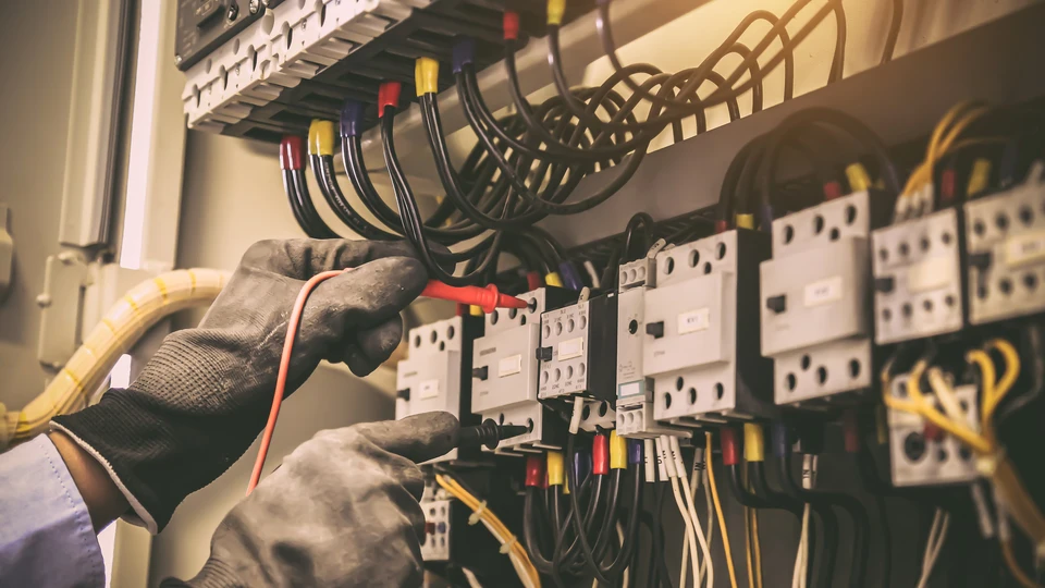 Électricien équipé de gants de protection travaillant sur un tableau électrique industriel, manipulant des câbles et contacteurs
