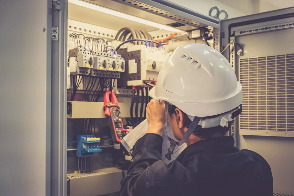 Électricien en casque blanc travaillant dans armoire électrique industrielle, utilisant multimètre rouge pour mesures